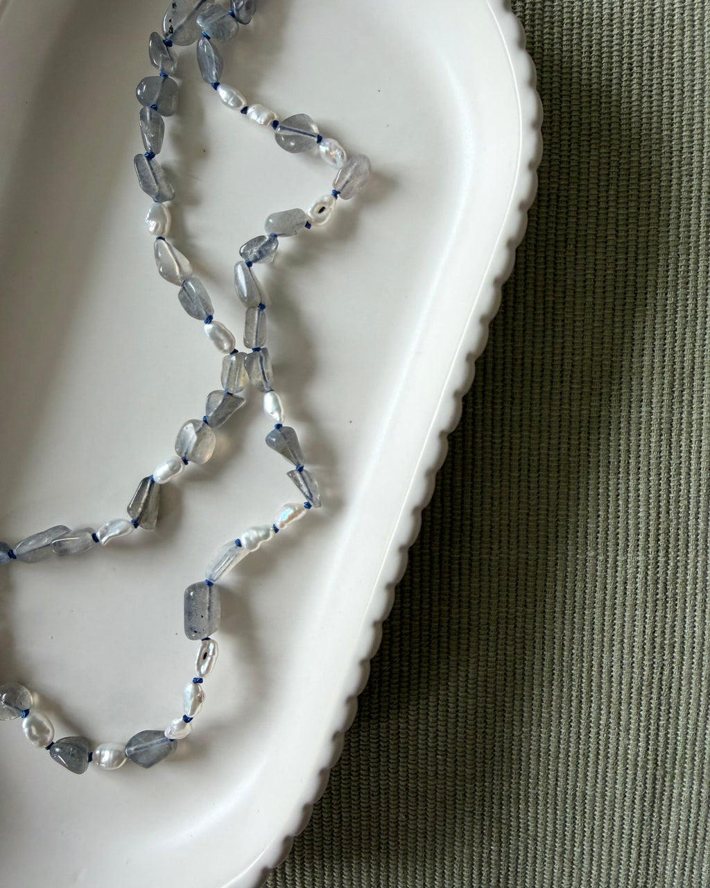 A beaded necklace made of labradorite, pearls and blue silk draped over a white scalloped ceramic dish on a textured olive green fabric background.