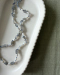 A beaded necklace made of labradorite, pearls and blue silk draped over a white scalloped ceramic dish on a textured olive green fabric background.