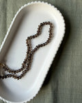A strand of dark brown translucent smoky quartz gemstone beads, draped inside a white ceramic dish on a textured olive green fabric background.
