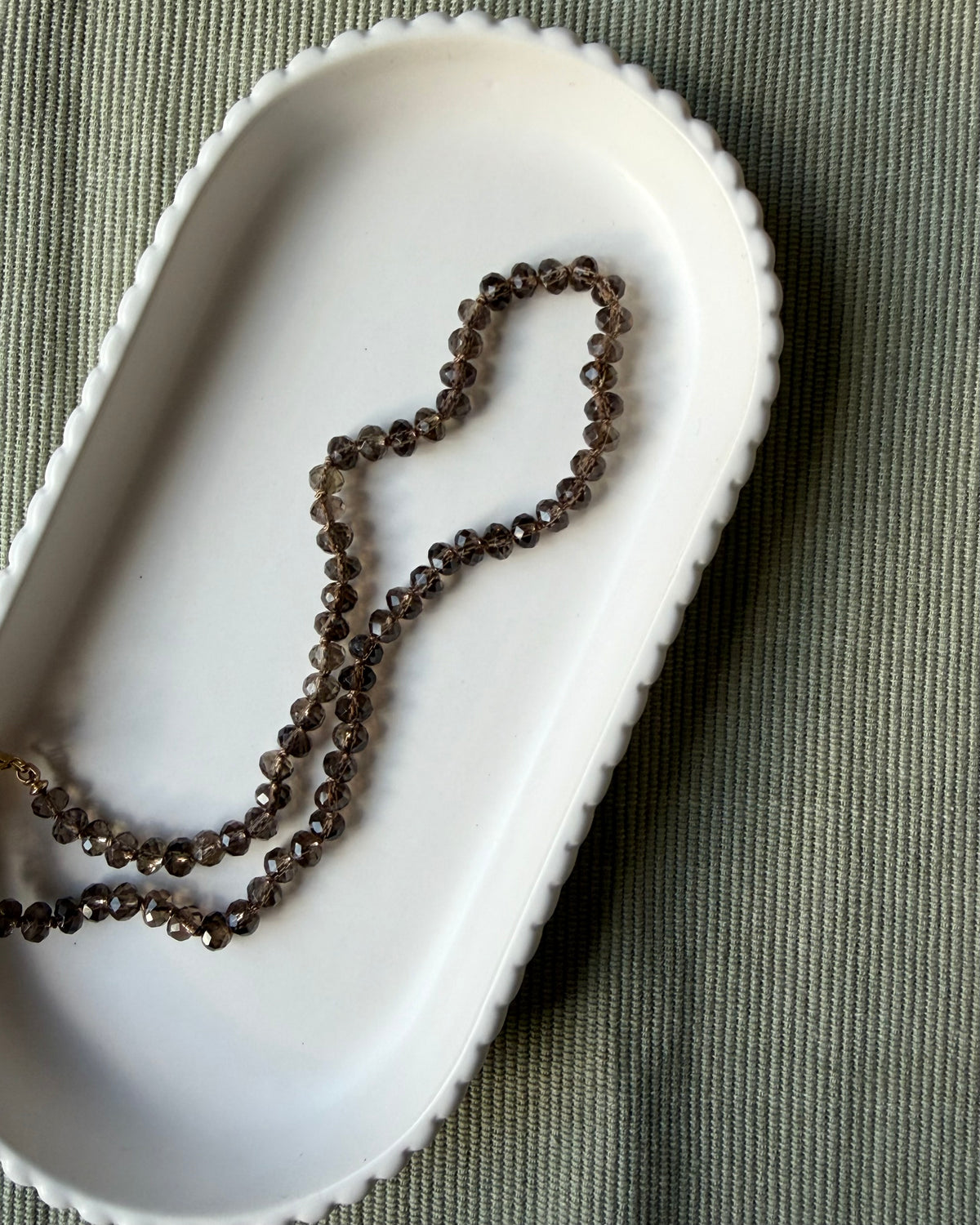 A strand of dark brown translucent smoky quartz gemstone beads, draped inside a white ceramic dish on a textured olive green fabric background.