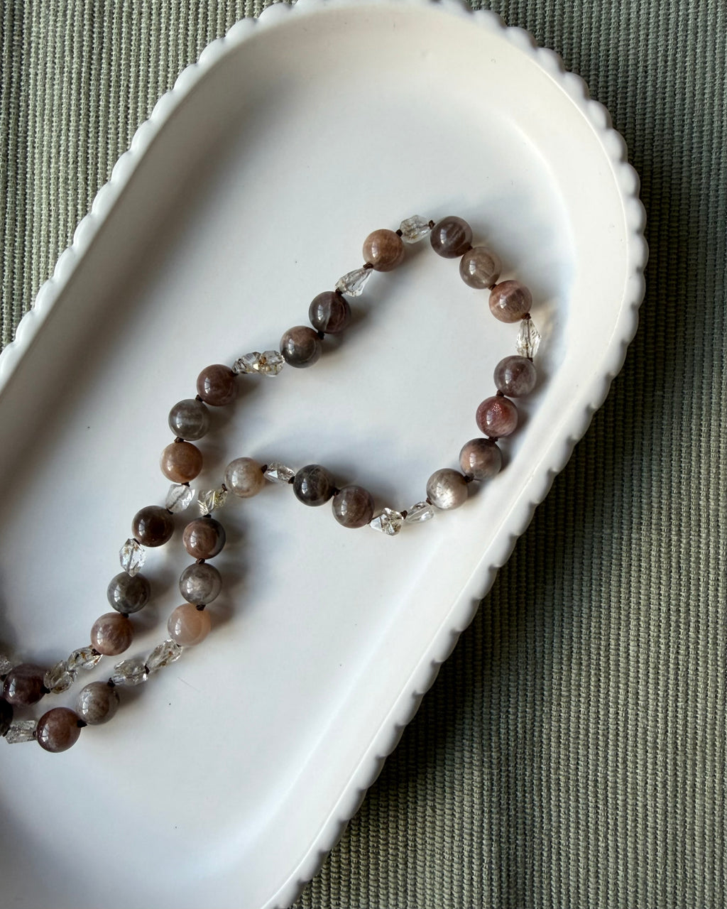 A beaded necklace with round brown moonstone gemstone beads and small clear crystal quartz accents, arranged in an oval white dish on a textured olive green fabric background.