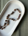 A beaded necklace with round brown moonstone gemstone beads and small clear crystal quartz accents, arranged in an oval white dish on a textured olive green fabric background.