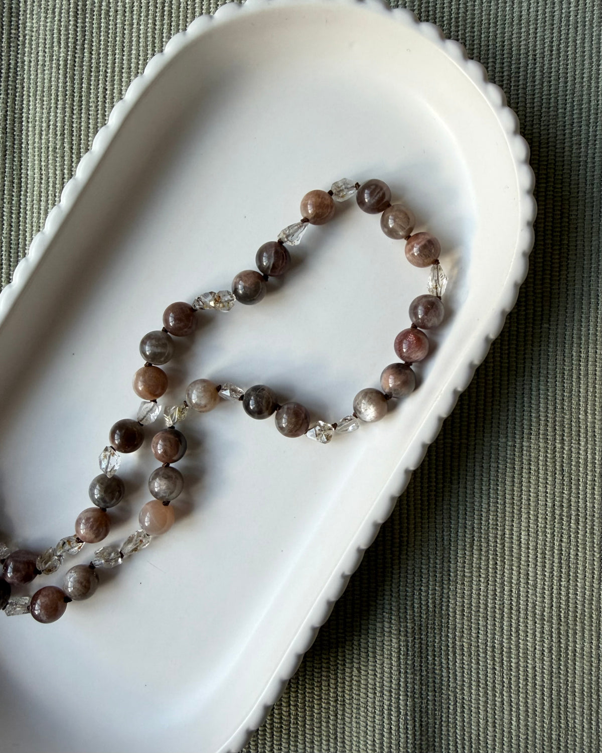 A beaded necklace with round brown moonstone gemstone beads and small clear crystal quartz accents, arranged in an oval white dish on a textured olive green fabric background.