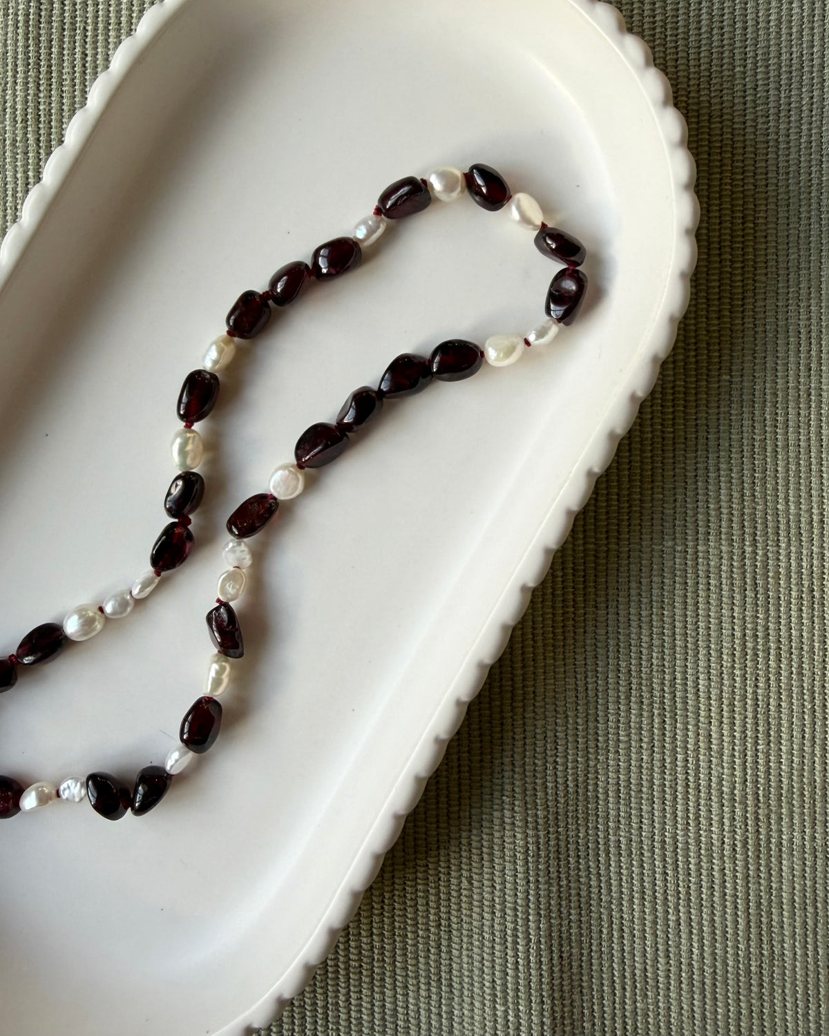 Dark red garnet nugget beads and small baroque pearls strung on a wine coloured silk, draped in a white scalloped dish on an olive background