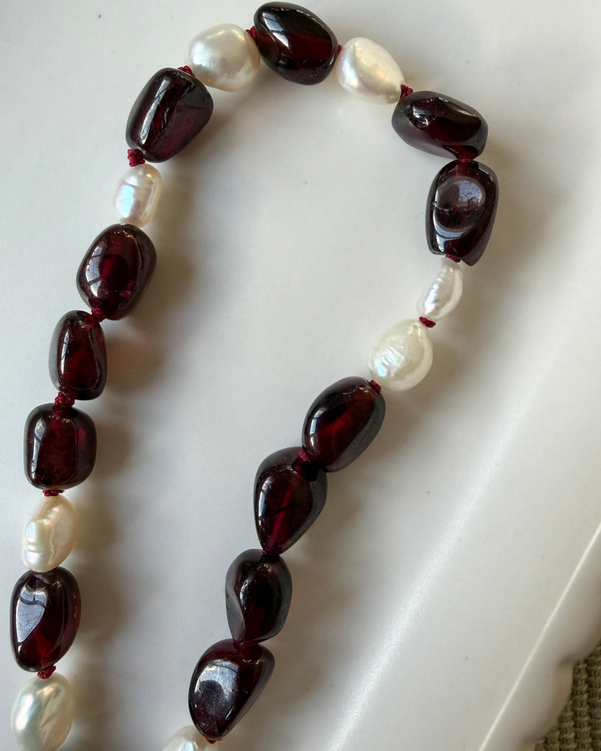 Dark red garnet nugget beads and small baroque pearls strung on a wine coloured silk, draped in a white scalloped dish on an olive background