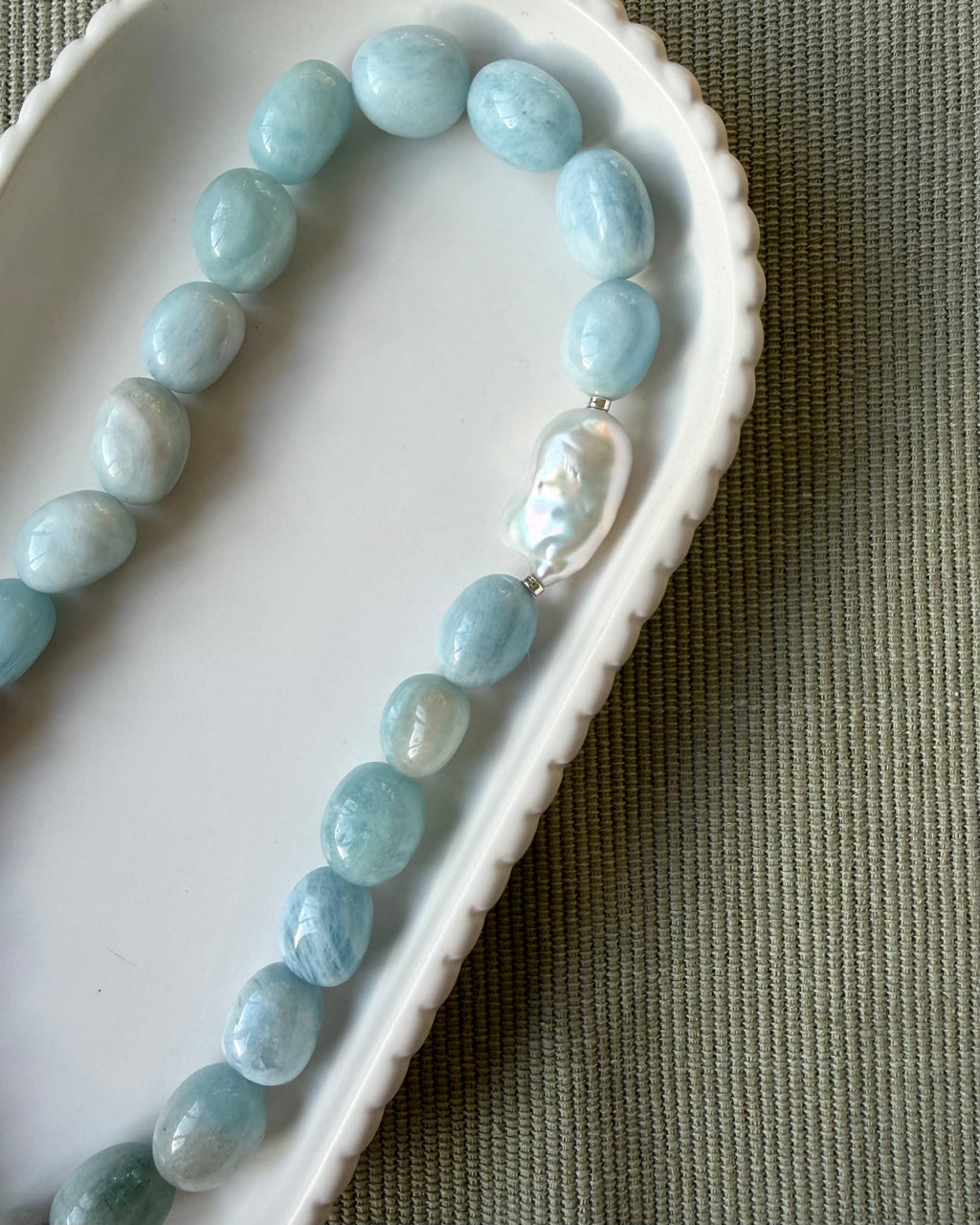 Large nugget aquamarine beads with a singular large baroque pearl on a scalloped white dish on an olive background