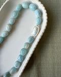 Large nugget aquamarine beads with a singular large baroque pearl on a scalloped white dish on an olive background
